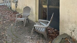 A two-story building with a beige exterior and large windows. There are two outdoor chairs and a table on a paved area with fallen leaves.