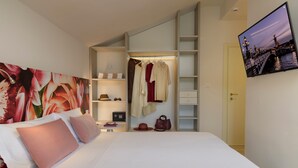 A hotel room with a bed, a large floral artwork on the wall, a television, and a wardrobe with hanging clothes. - Albergo Roma (Gatteo a Mare)