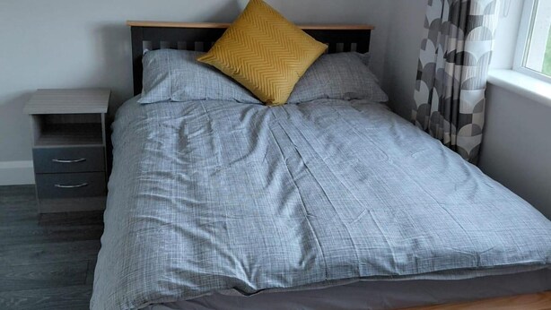 Ein Bett mit grauer Bettdecke und zwei Kissen, eines gelb und eines grau, in einem Zimmer mit Holz-Kopfteil, einem Nachttisch mit Schubladen und einem Fenster mit Vorhängen.
