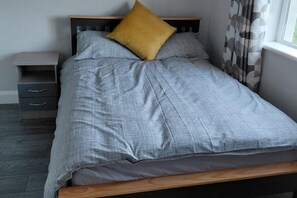 A bed with a grey bedspread and two pillows, one yellow and one grey, in a room with a wooden headboard, a nightstand with drawers, and a window with curtains. - 4 Bedroom House Sleeps 8 -pets Welcome -fireplace (Galway)