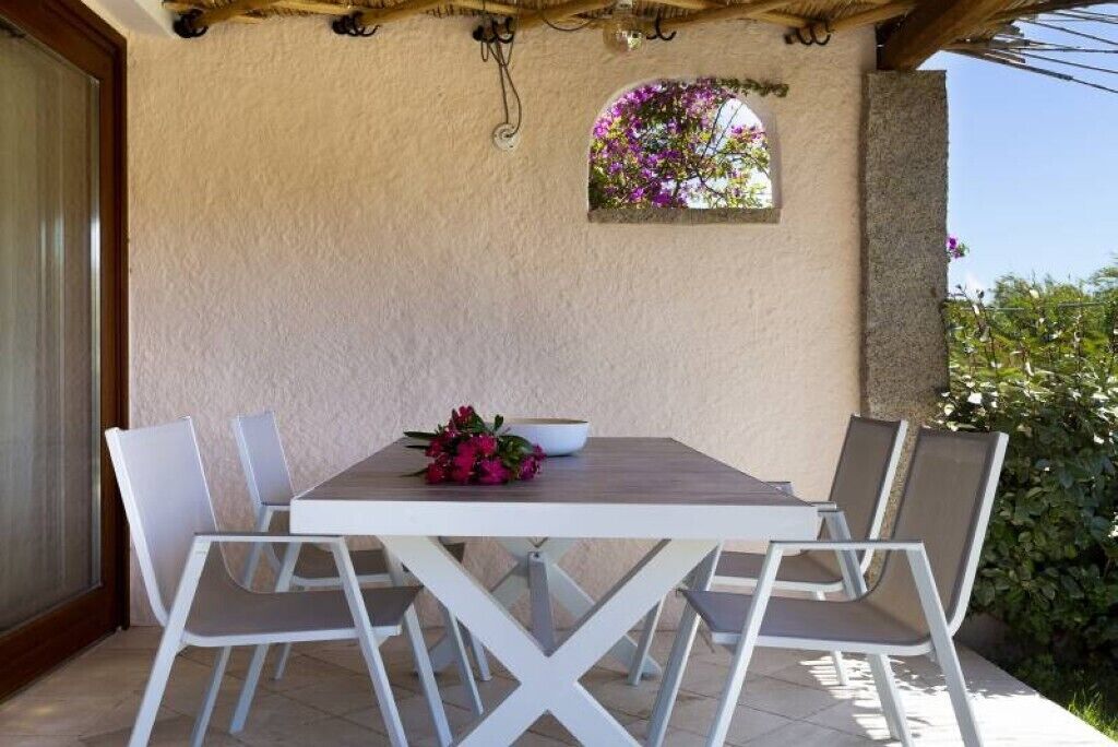 An outdoor patio with a white table and four chairs, a small decorative window, and a wall-mounted light fixture.
