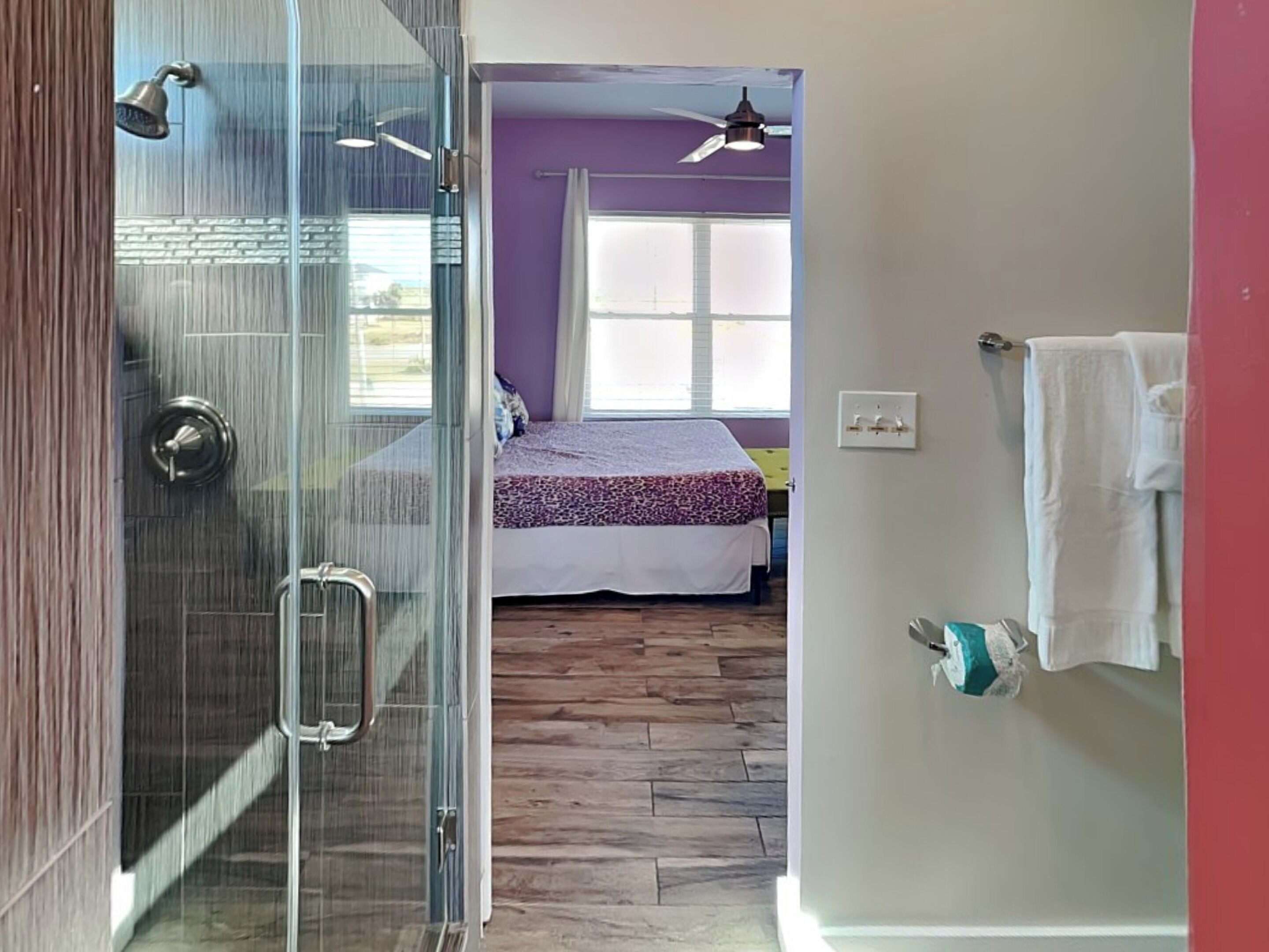 A room with a glass shower, a bed with a patterned bedspread, a ceiling fan, and a window with curtains.