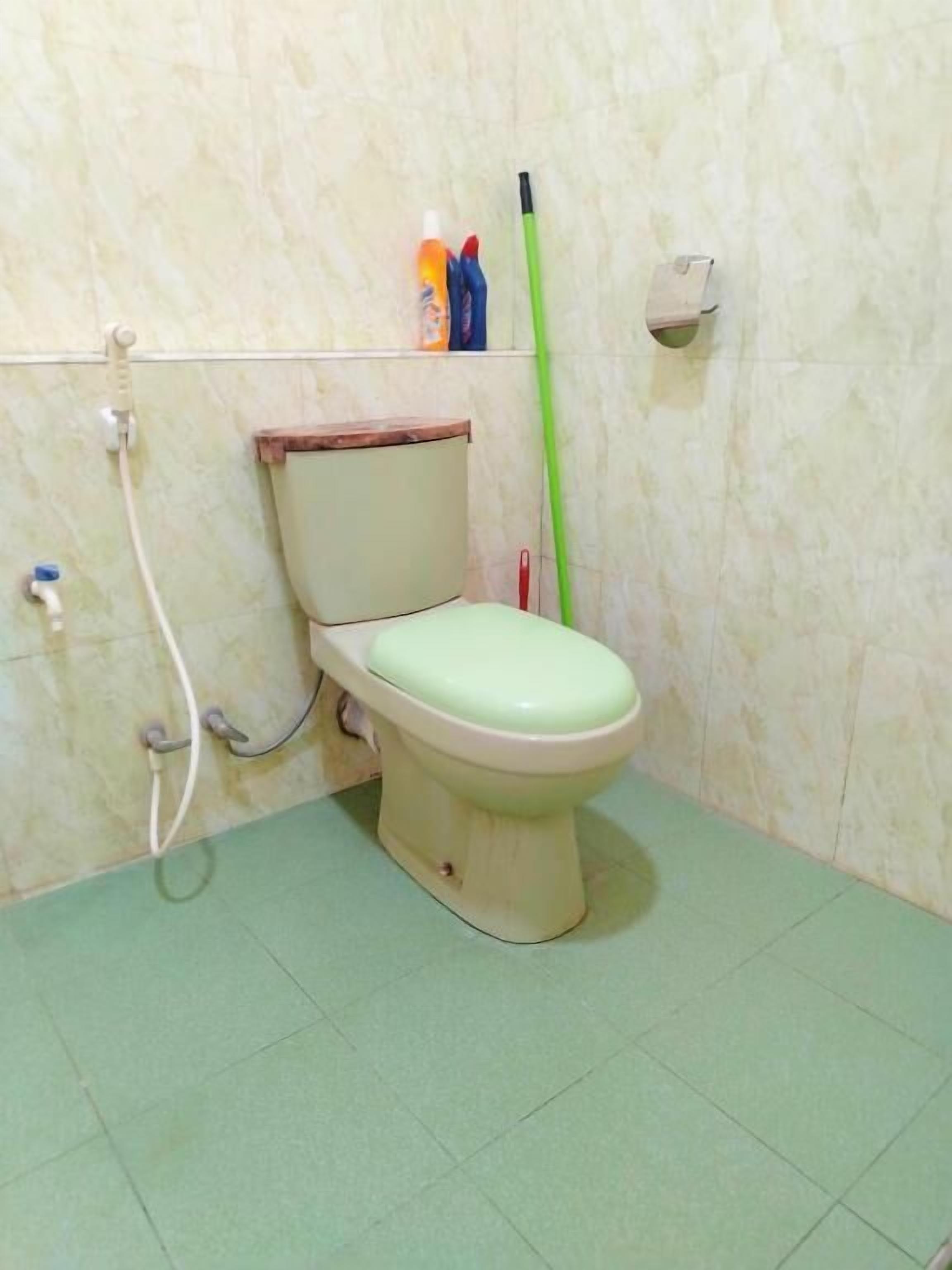 Een toilet met een groen zitje en een houten deksel, een groene bezem die tegen de muur staat, en een handdroger aan de muur.