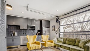 A modern kitchen with grey cabinets, a microwave, and a refrigerator. There is a yellow armchair and a green sofa in the living area. - Downtown Condo · Walk to Broadway · Free Parking (Nashville)