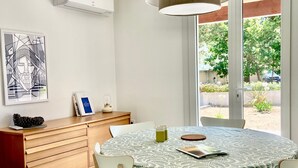 Un comedor con una mesa redonda, sillas, mantel estampado, lámpara colgante, aire acondicionado de pared y una puerta que da a un área exterior.