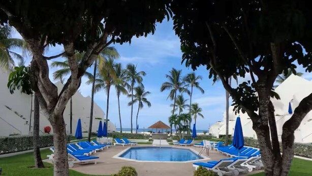 Zona de piscina con sillones de descanso y sombrillas, rodeada de palmeras y un cielo despejado.