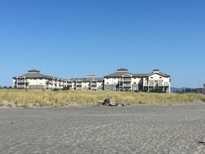 Een strandhotel met meerdere verdiepingen en balkons, gelegen aan een zanderig strand met hoge grassen op de voorgrond.