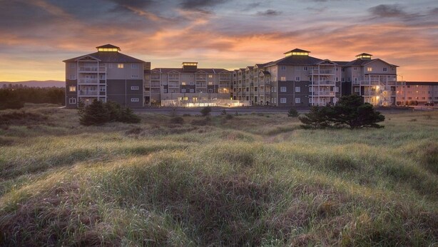 A multi-story residential building with a flat roof and multiple balconies, surrounded by grassy fields and a sunset sky. - Long Beach WA | Coastal Family Retreat | Pool + Hot Tub | 2BR (Long Beach)