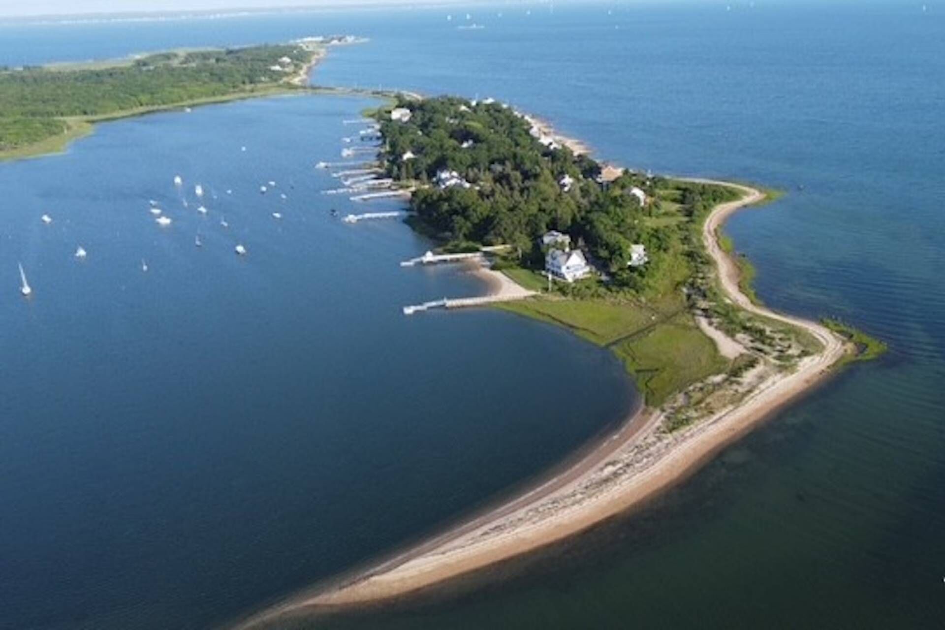 Vue aérienne d’une zone côtière avec une petite île comprenant quelques bâtiments, une plage de sable et un port de plaisance avec des bateaux.