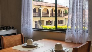 Una mesa de comedor con individuales y una vela, una ventana con cortinas de encaje, y vista a un edificio con arcos.