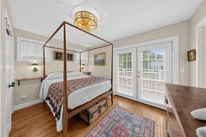 A bedroom with a four-poster bed, a wooden chest of drawers, a patterned rug, and a large window with blinds.