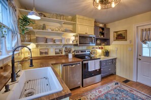 Una cocina con armarios de madera, lavavajillas de acero inoxidable, microondas y estufa. Se ven estantes con vajilla y una planta a la izquierda.