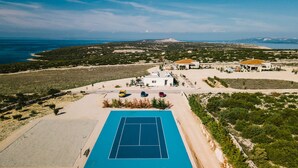 En vy från ovan av en fastighet vid kusten, med en tennisbana, flera byggnader och utsikt över havet och kullarna.