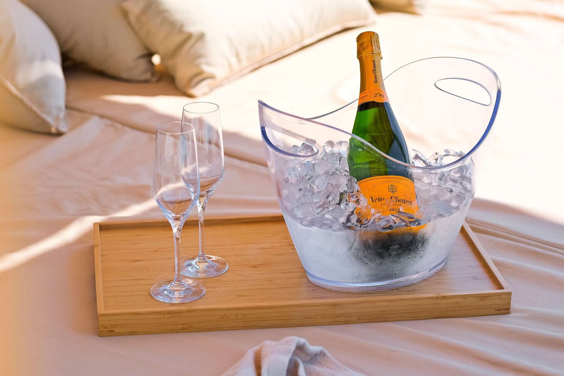 Een houten dienblad met een champagnefles in een glazen schaal vol ijs, twee lege glaasjes, en een bed met kussens op de achtergrond.