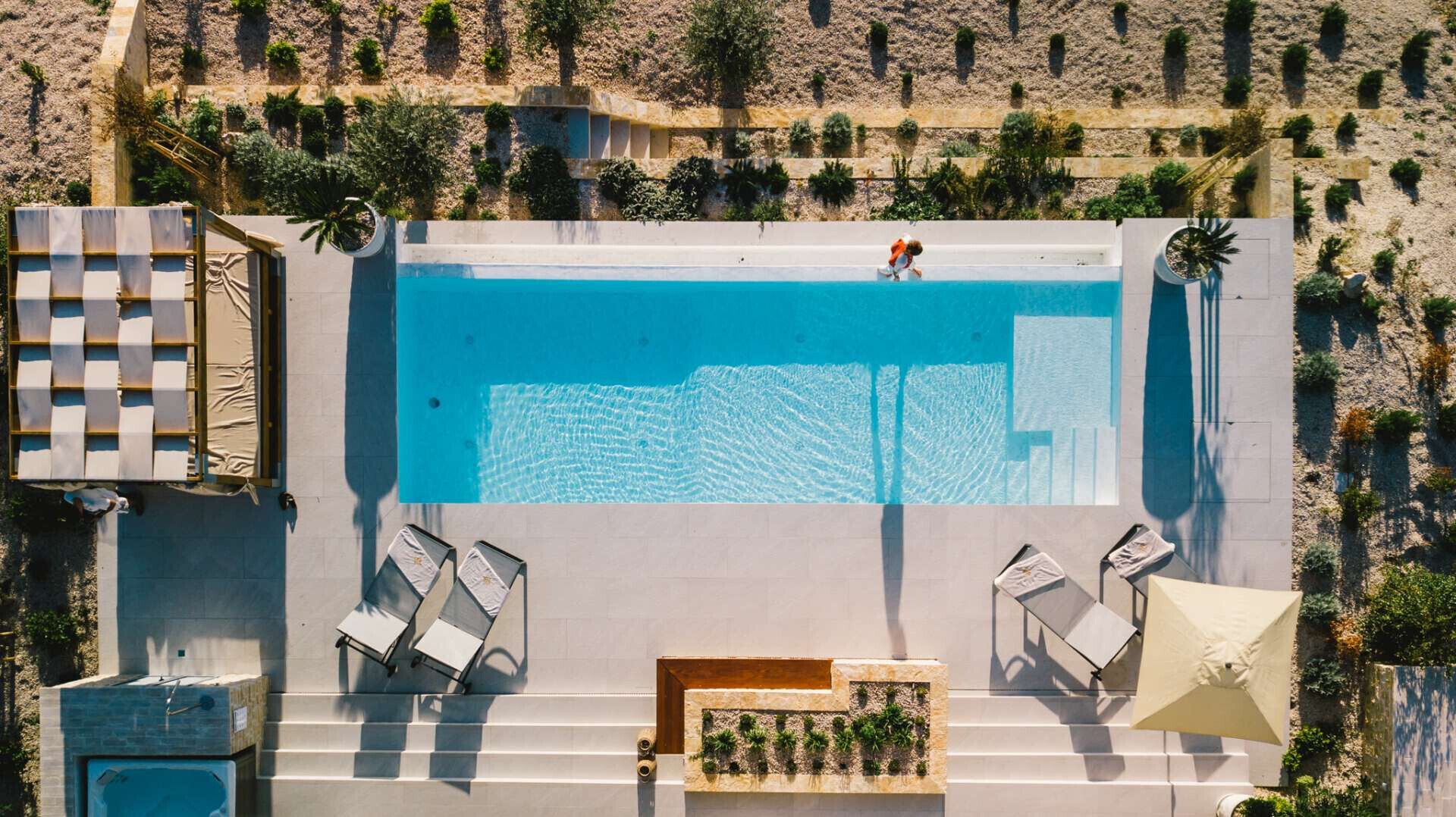 An aerial view of a rectangular swimming pool with a clear blue surface, surrounded by a stone wall and a landscaped area with small plants. There are two lounge chairs and a sun umbrella on the poolside.