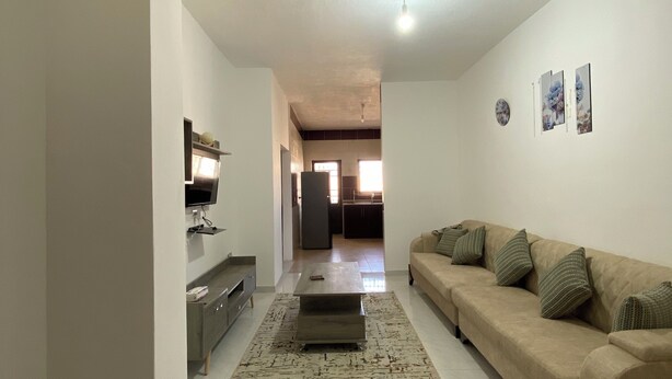 Amplia sala de estar con un sofá color beige, una mesa de centro y un televisor de pantalla plana sobre un mueble. Se observa una alfombra estampada en el suelo, paredes blancas y piso de baldosas.