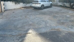 Un SUV blanco estacionado en un área de estacionamiento de concreto, con un muro blanco y algo de vegetación al fondo.