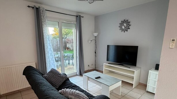 Una sala de estar con un sofá negro, una mesa de centro blanca, un televisor sobre un mueble bajo y una puerta corrediza de vidrio que da a un área exterior.