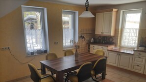 A dining area with a wooden table and chairs, a vase with dried plants, and a kitchen with white cabinets and a window.