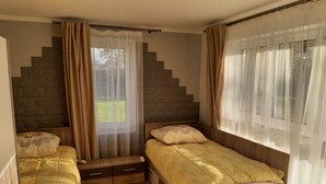 A bedroom with two single beds, a small window with sheer curtains, a wooden headboard, and a stone wall behind the window.