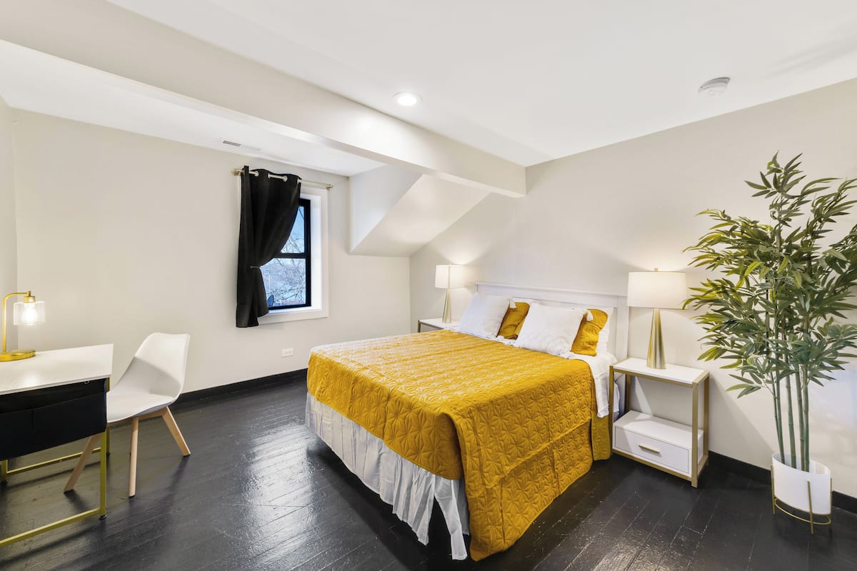 A modern hotel room with a large bed, a desk, a chair, and a potted plant.