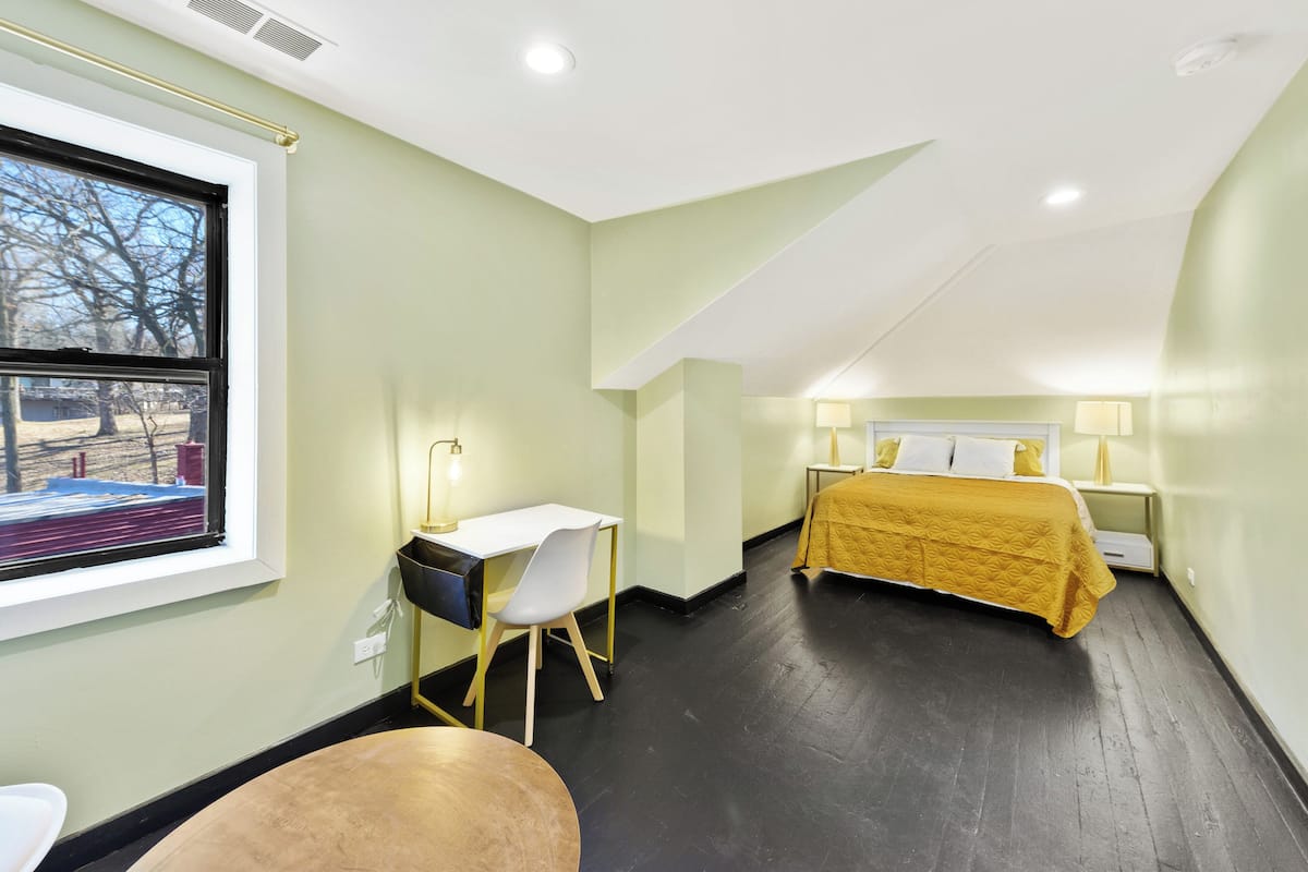 A bedroom with a bed, bedside table, lamp, and a desk with a chair.