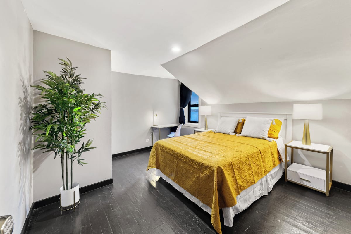 A modern bedroom with a large bed, a bedside table, a nightstand, a potted plant, and a window.
