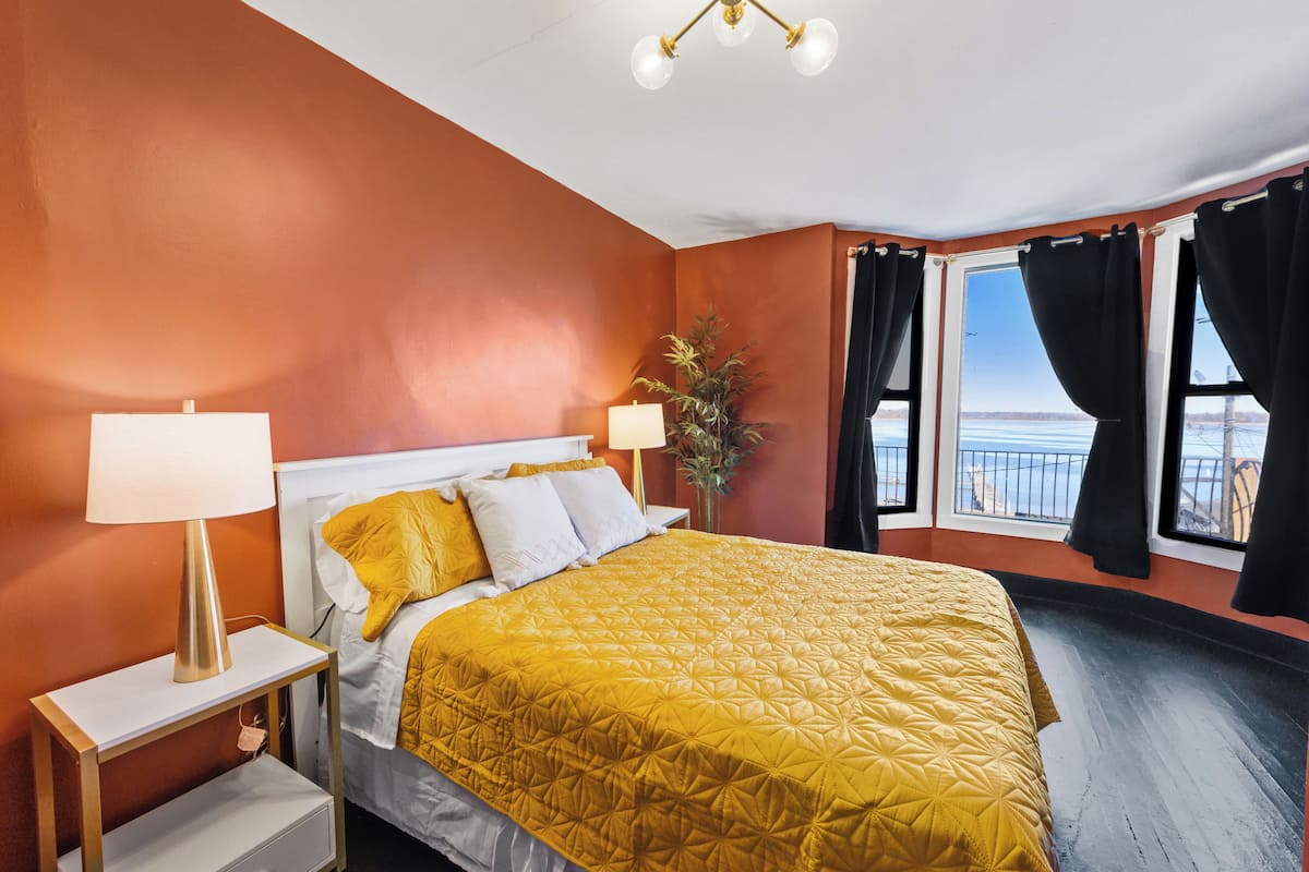 A hotel room with a large bed, two bedside lamps, a nightstand, a potted plant, and a view of the ocean through the windows.