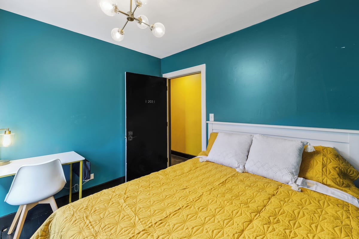 A room with teal walls, a yellow bedspread, a white desk, and a black wardrobe.