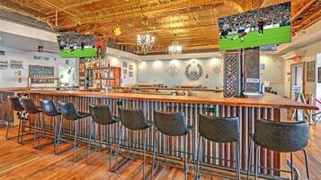 A bar area with a long counter, multiple stools, and a television screen displaying a sports event.