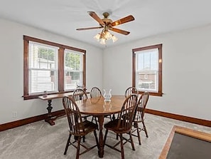 Une salle à manger avec une table ronde, six chaises, un ventilateur de plafond et deux fenêtres équipées de stores.