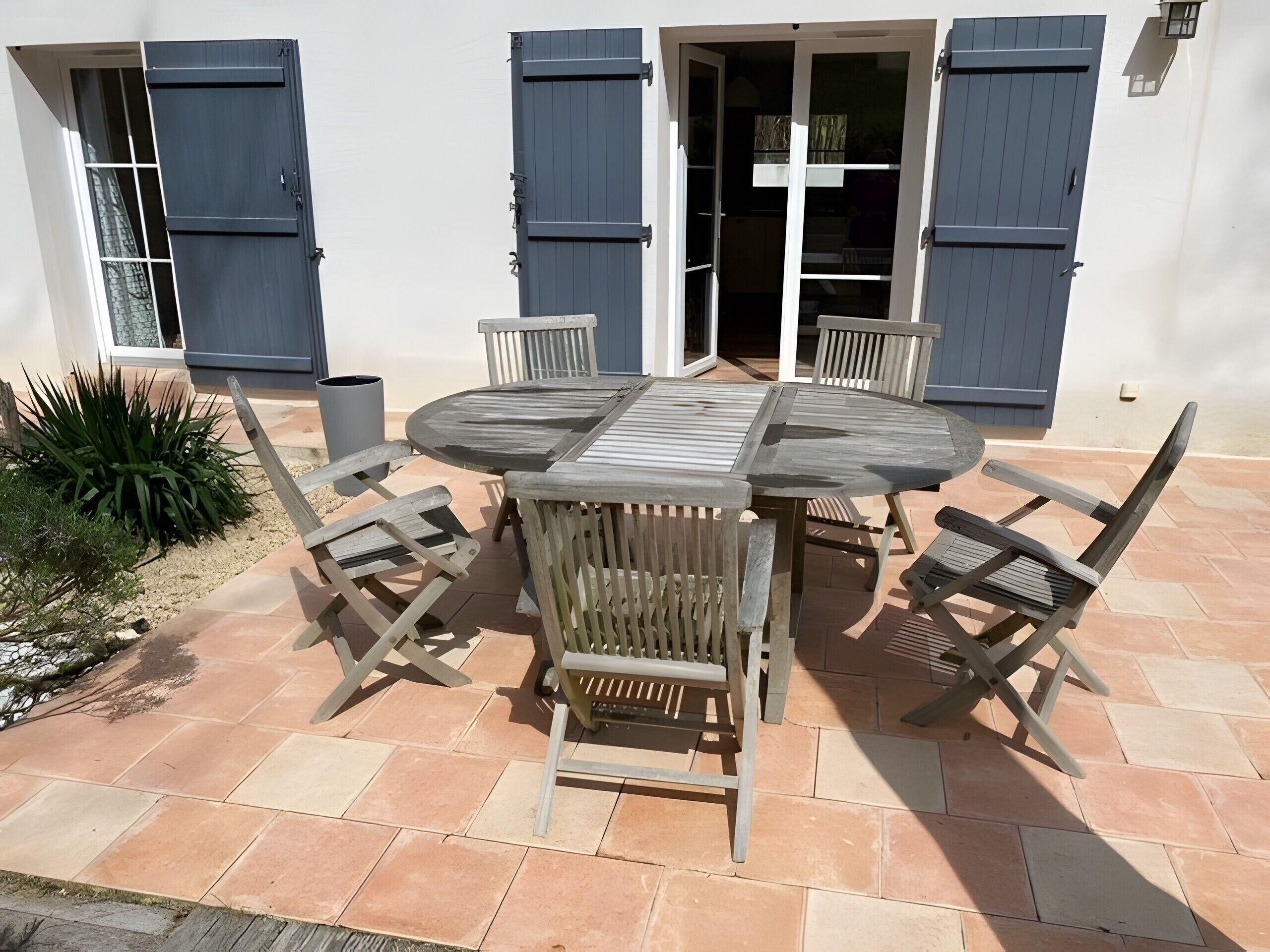 Un patio al aire libre con una mesa de madera redonda y seis sillas, rodeado de un suelo de baldosas y un pequeño jardín.