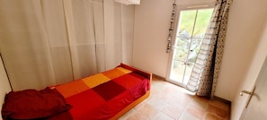 A single bed with a red and orange bedspread in a room with a sliding door and a view of the outdoors.
