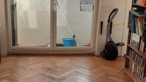 Et værelse med fiskebensparket gulv, en skydedør til en balkon, en reol og en støvsuger.