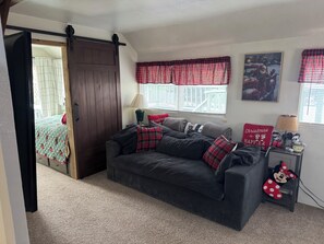 Ein gemütliches Wohnzimmer mit einem dunklen Sofa, roten und schwarzen Karokissen, einem kleinen Tisch mit einer Lampe und einer Minnie Maus Dekoration.