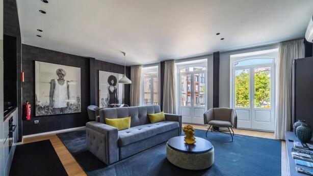 Ein modernes Wohnzimmer mit grauem Sofa, einem runden Couchtisch und einem Sessel. Große Fenster mit Vorhängen, ein Kamin und Kunstwerke an den Wänden.