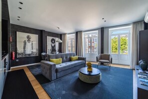 Una sala de estar moderna con un sofá gris, una mesa de centro redonda y un sillón. Hay ventanas amplias con cortinas, una chimenea y obras de arte en las paredes.