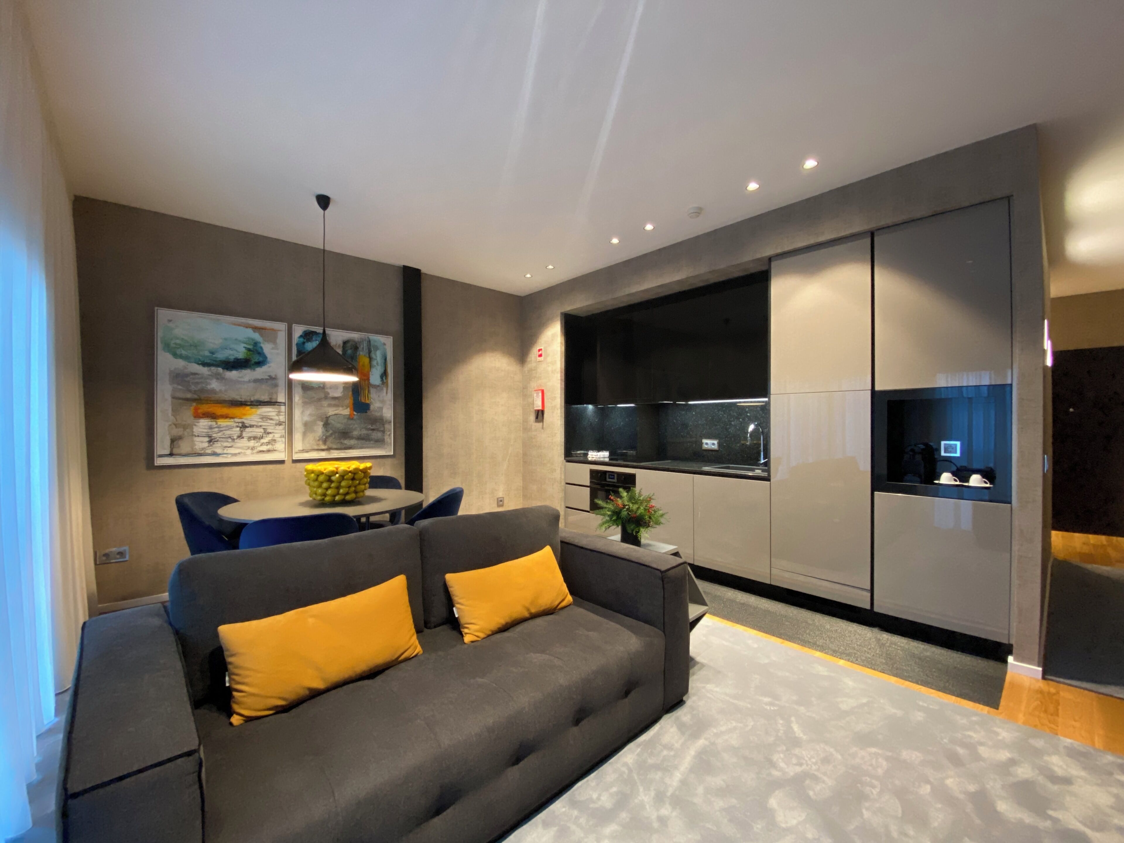 Een moderne woonkamer met een grijze bank, gele kussens, een eettafel met stoelen, een keuken met een zwart aanrecht en kunstwerken aan de muren.