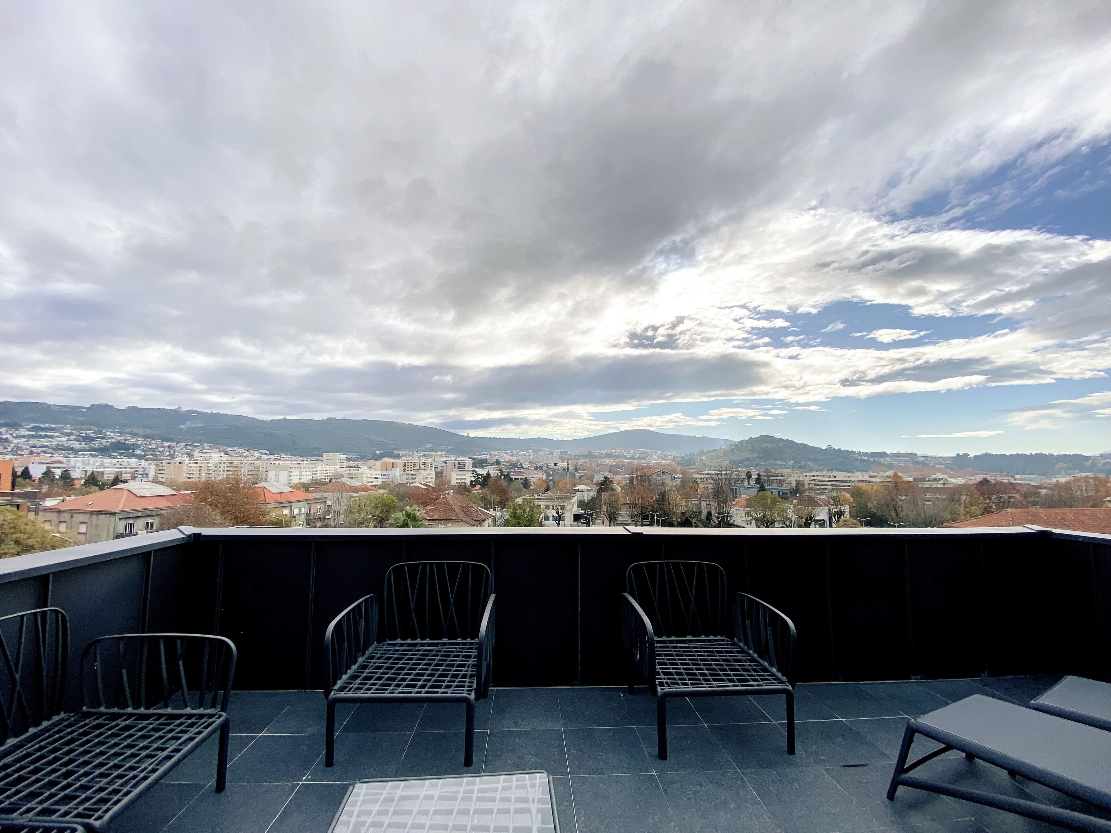 Terraza en la azotea con dos sillas de metal y una mesa, con vista a un paisaje urbano de edificios y colinas.