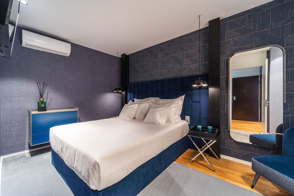 A modern bedroom with a large bed, a bedside table, a mirror, and a blue chair.