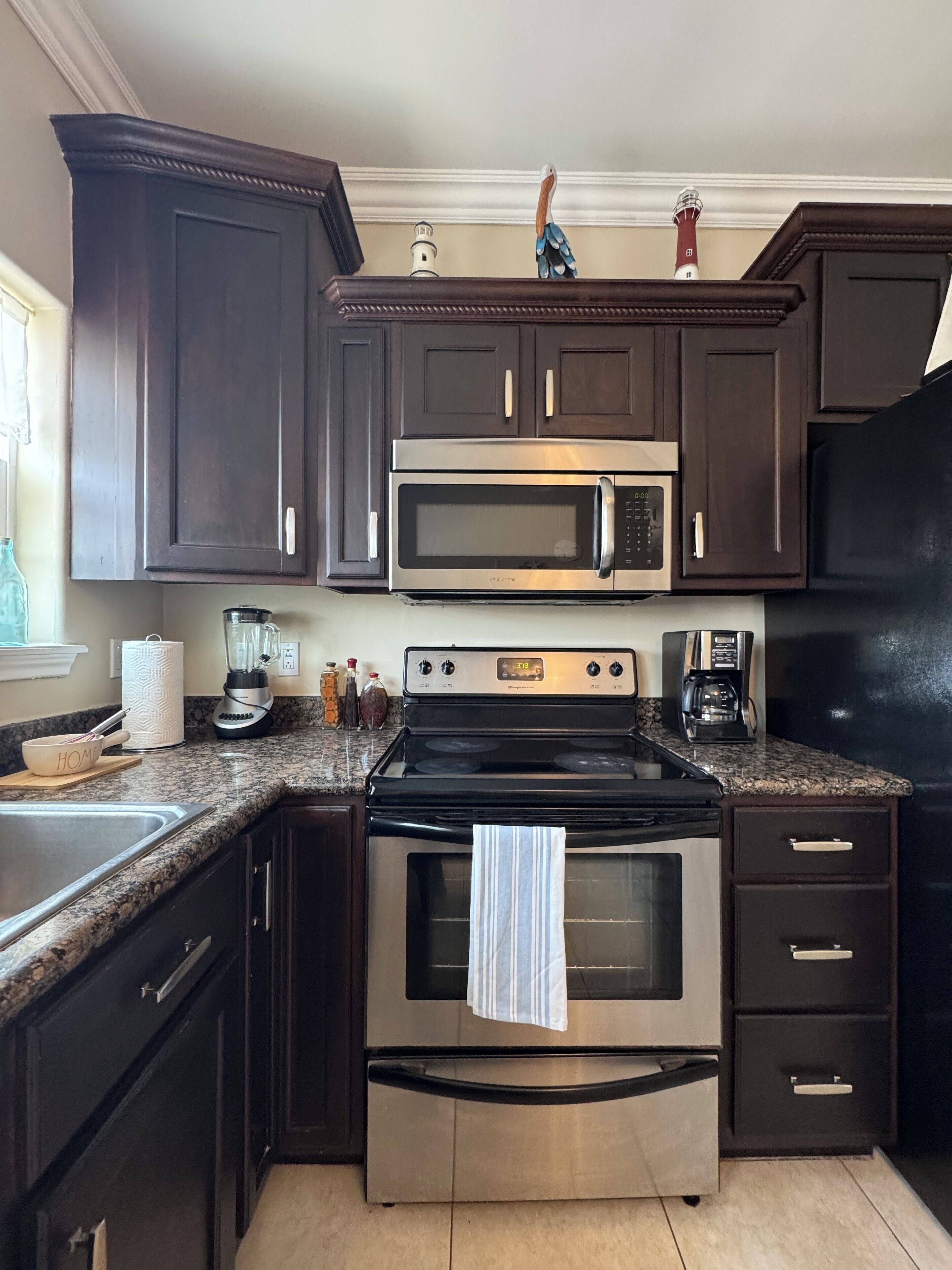 Una cocina con armarios de madera oscura, electrodomésticos de acero inoxidable, incluyendo un microondas, horno y refrigerador, una encimera de granito y un ventanal con vistas al exterior.