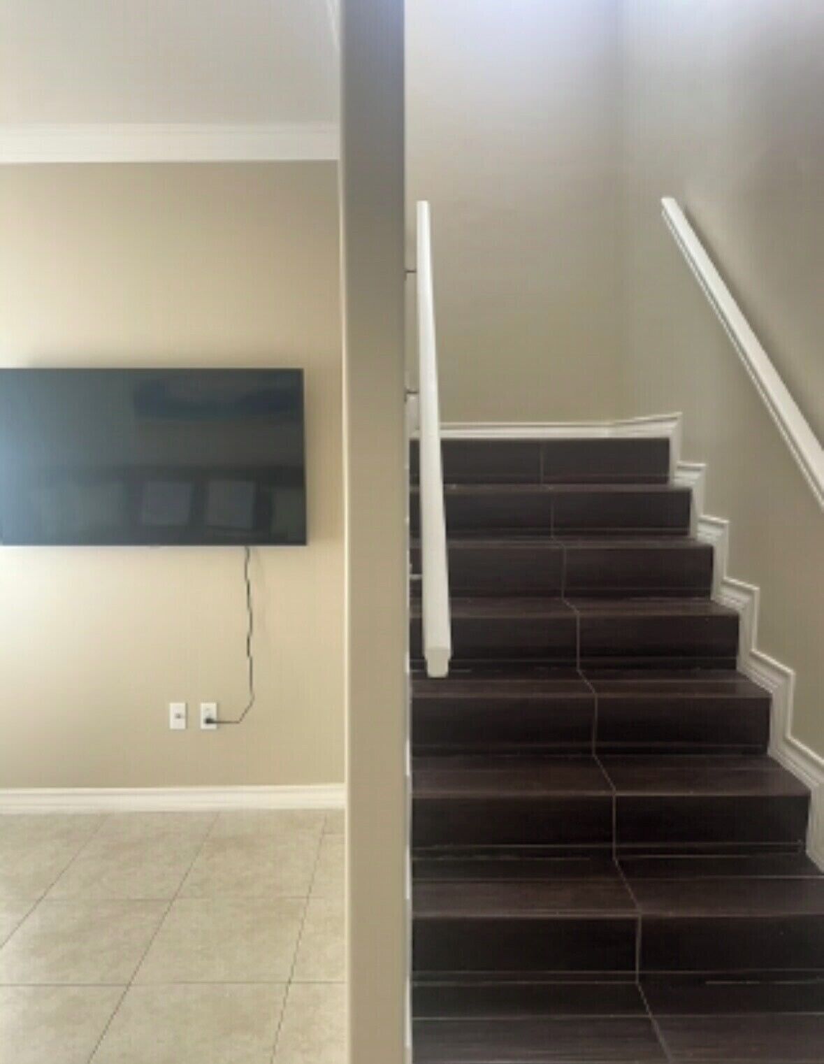 Un escalier aux marches sombres et aux rampes blanches, menant à un palier où est fixée un téléviseur à écran plat au mur.