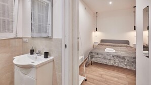 Un dormitorio moderno con cama, mesitas de noche, un tocador, una ventana con cortinas y un baño con lavabo y espejo.