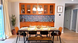 Uma sala de jantar com uma mesa de madeira posta para quatro pessoas, rodeada de cadeiras, com armários de madeira e painel decorativo.