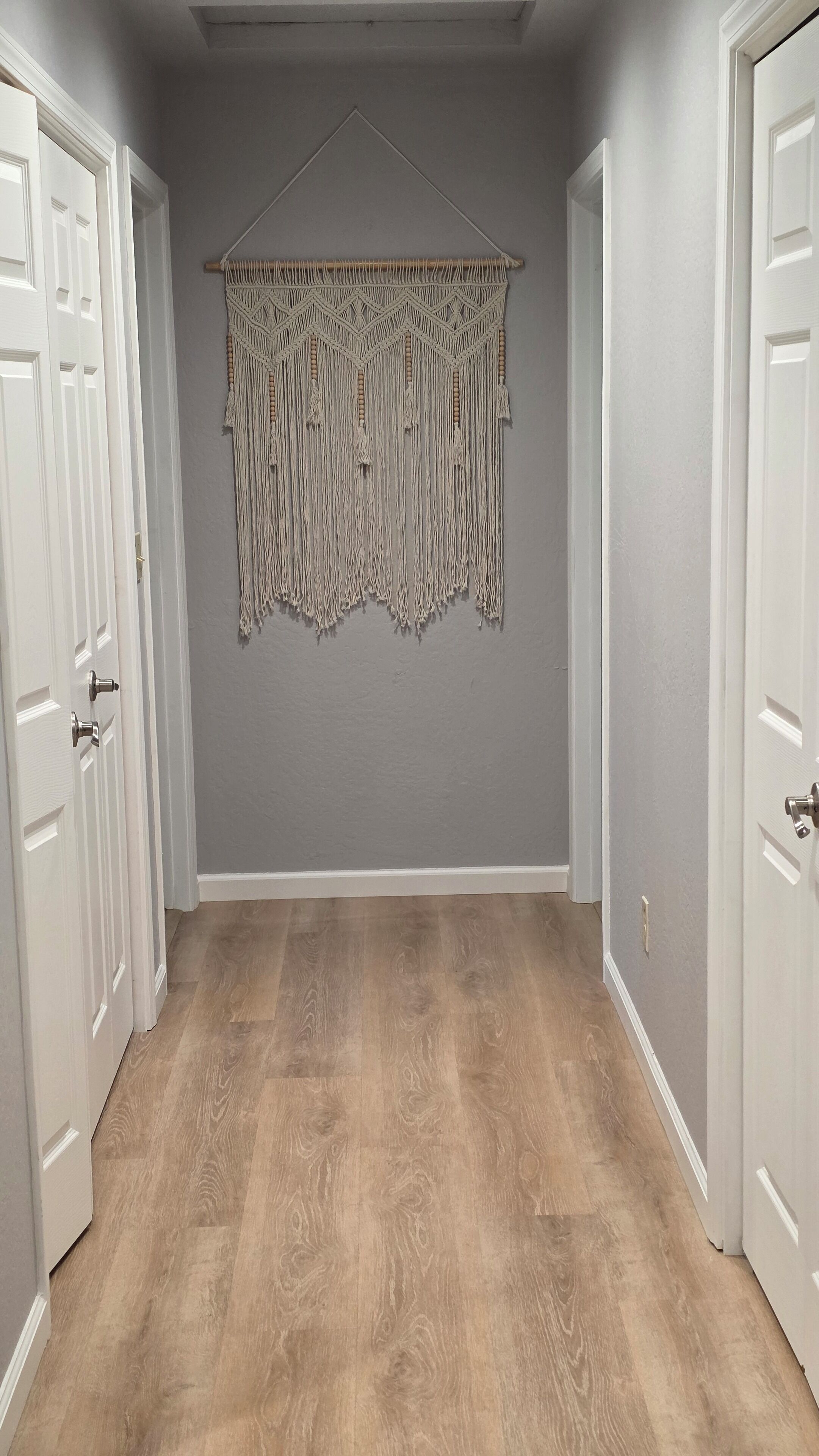 Een gang met houten vloer, grijze wanden en een macramé wanddecoratie.