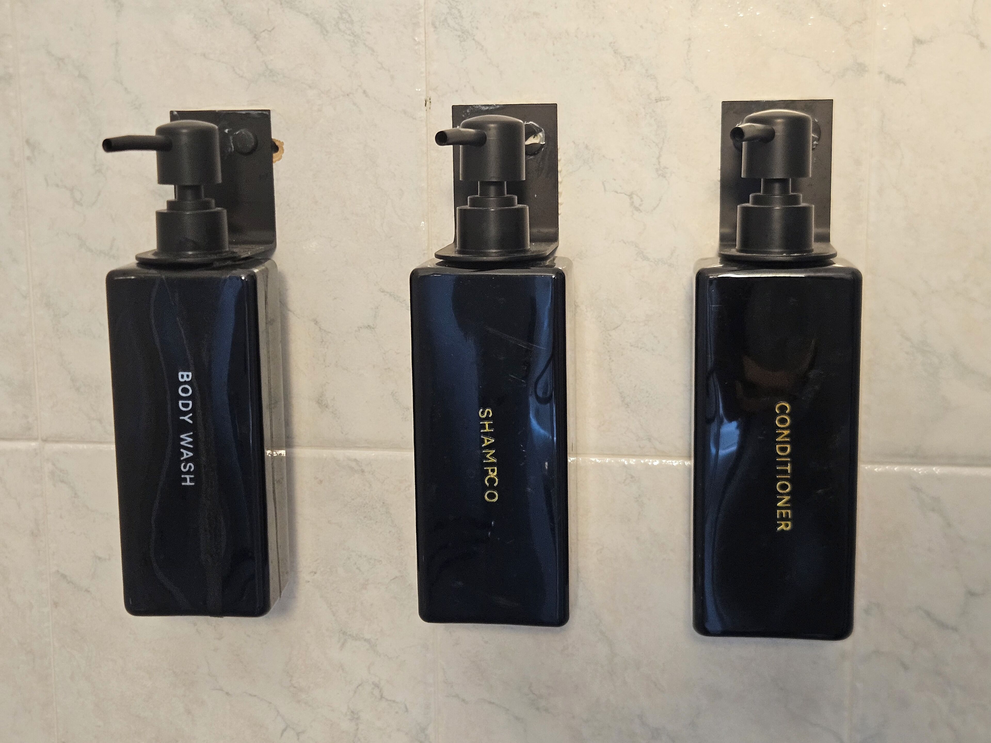 Drei schwarze Flaschen mit Dosierern, beschriftet mit 'BODY WASH', 'SHAMPOO' und 'CONDITIONER', vor einer weißen, gefliesten Wand.