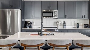 A modern kitchen with grey cabinets, a stainless steel refrigerator, a microwave, and a gas stove. There are three bar stools in front of the counter. - Cozy Fall Retreat*Near Dollywood/Island*HotTub*Xbox (Pigeon Forge)