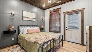 A bedroom with a bed, a nightstand, a wall-mounted lamp, a window with blinds, and a door. - Cozy Fall Retreat*Near Dollywood/Island*HotTub*Xbox (Pigeon Forge)