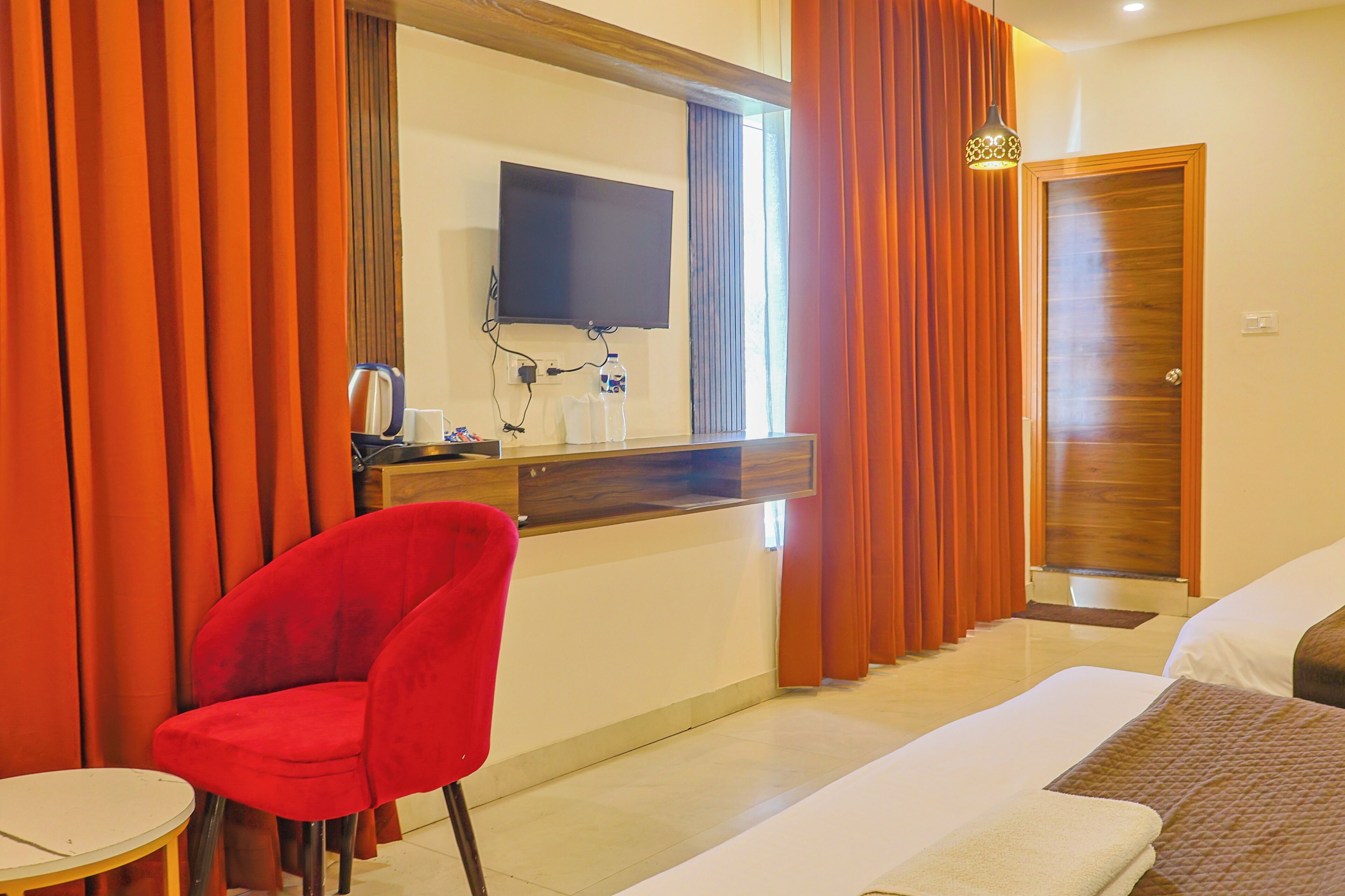 A hotel room with a bed, a red chair, a small table, a TV mounted on the wall, and a wooden door.