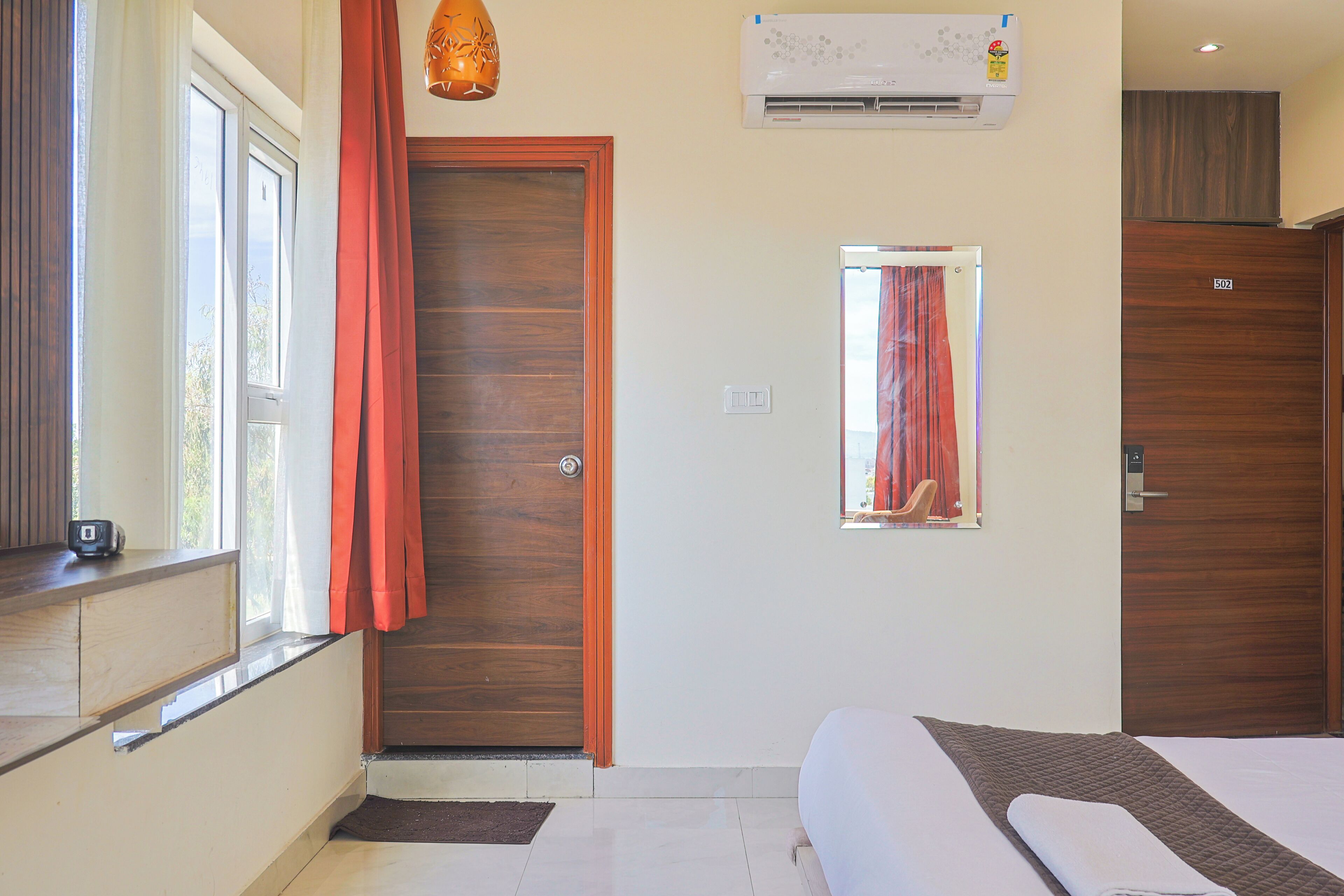 A bedroom with a bed, a window with curtains, a door, an air conditioning unit, and a small mirror.